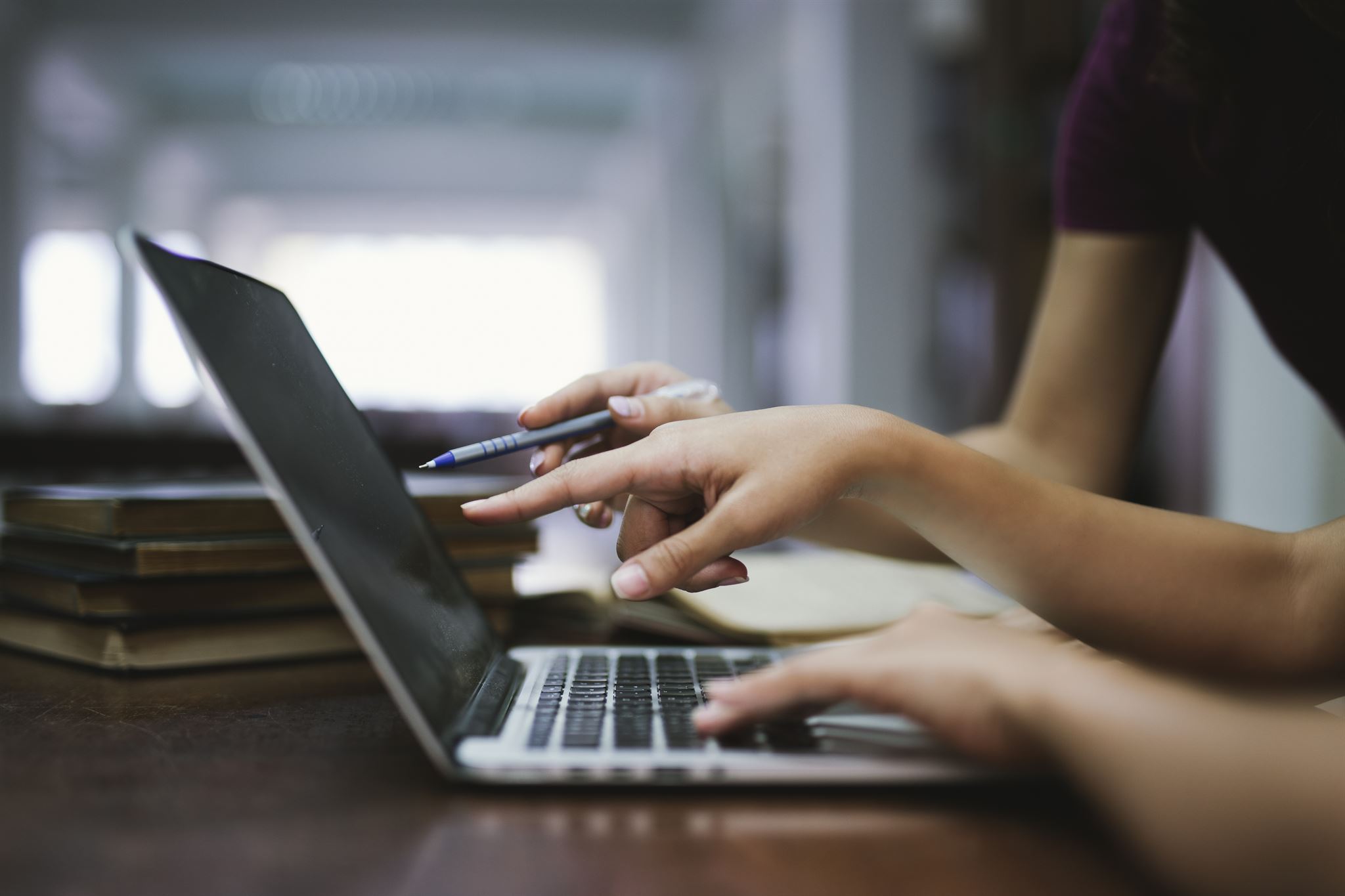 Close-up-of-two-peoples-hands-pointing-at-laptop-screen-with-stack-of-4-books-in-background,-one-person-holding-a-pen